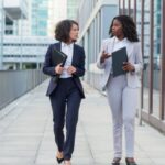 Two professional women walking and talking outdoors in business attire.