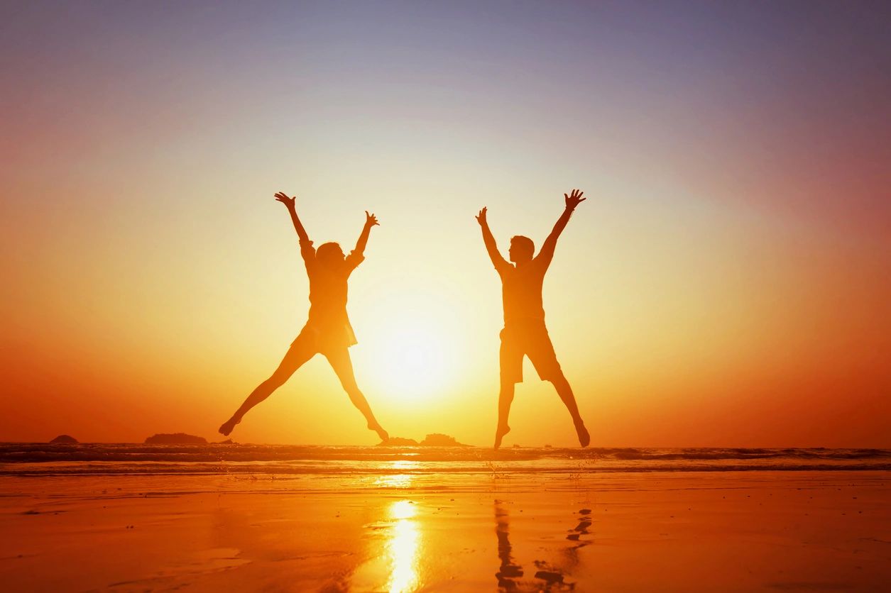 Two people joyfully jump at sunset on a reflective beach.