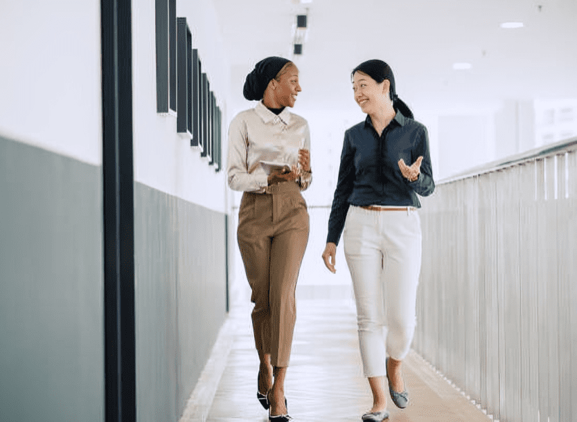 Two professional women walking and talking in a modern office corridor.